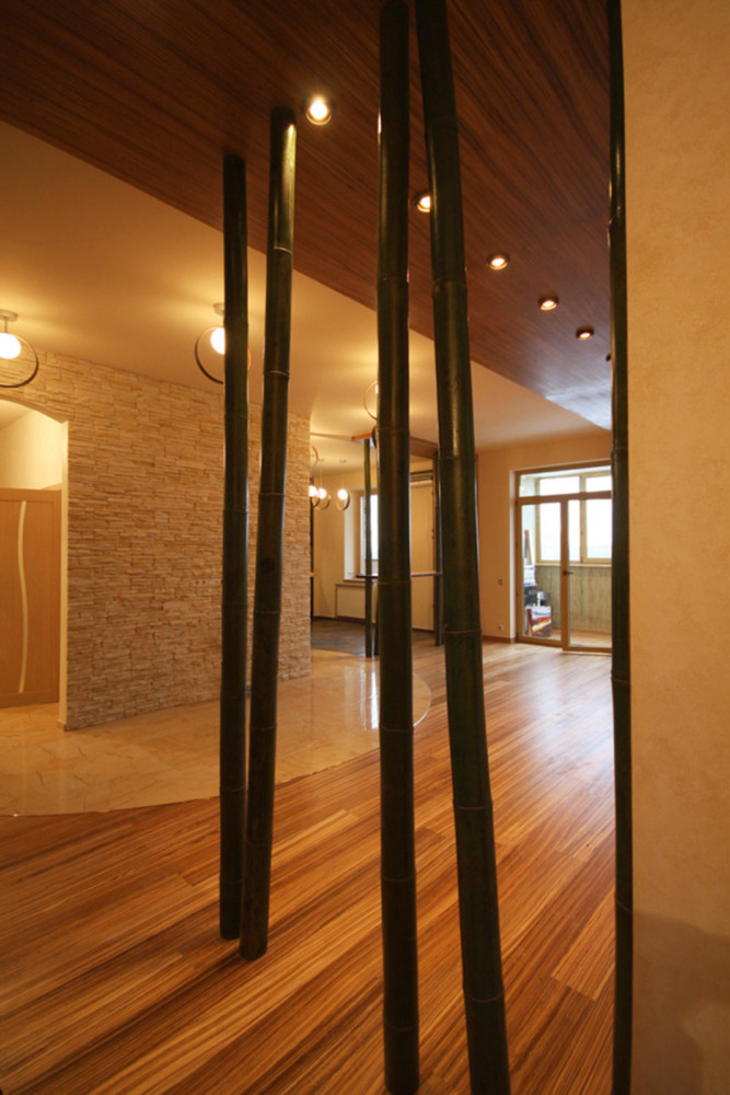 Деревянную тему дизайна поддерживает ряд колонн из бамбука. На белой части потолка контрастно работают современные круглые светильники. Из прихожей полукругом выдается зона на полу, облицованная керамической плиткой с каменной текстурой.